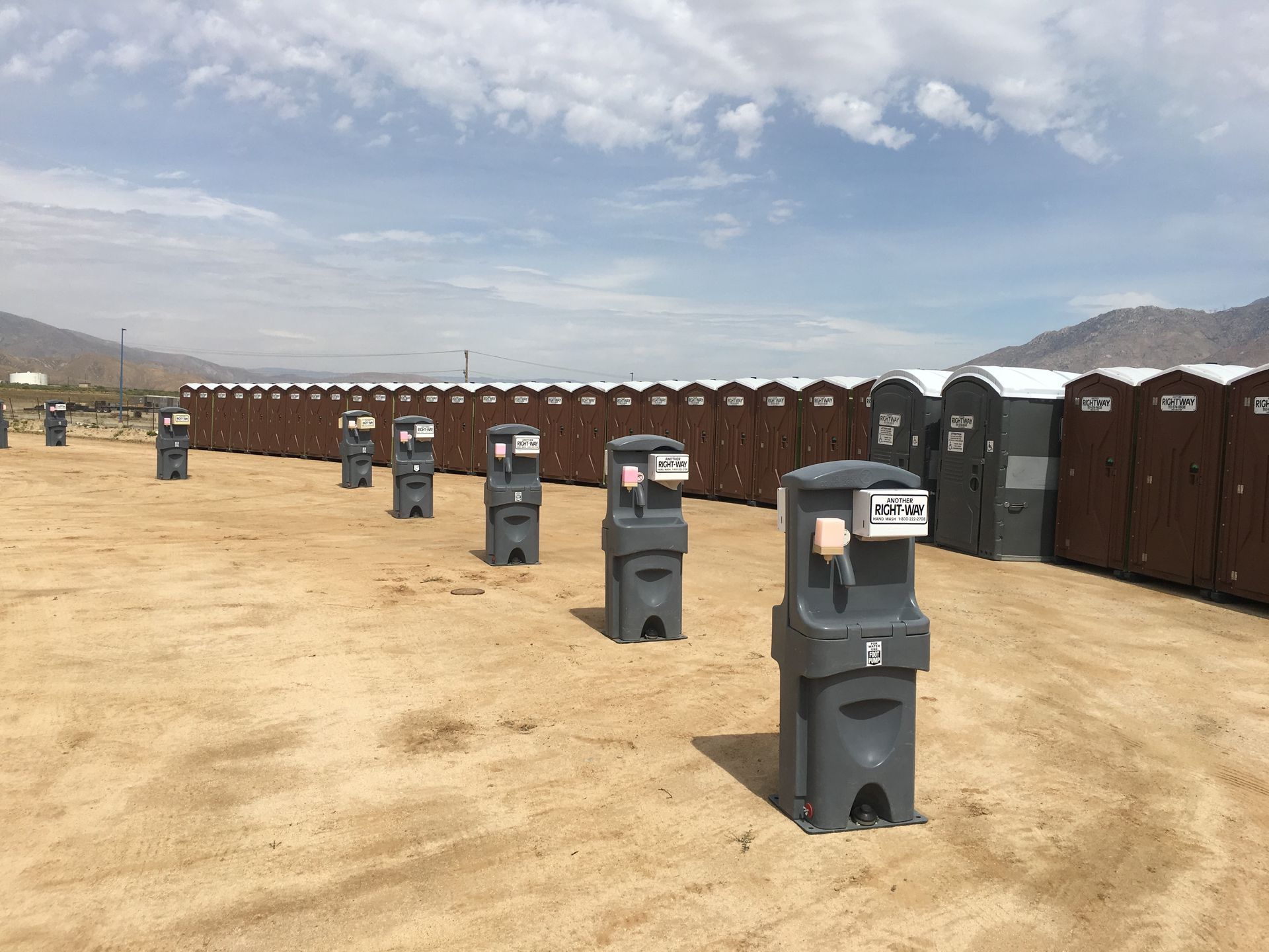 Rows of portable toilets and hand washing stations in a desert landscape under a cloudy sky.