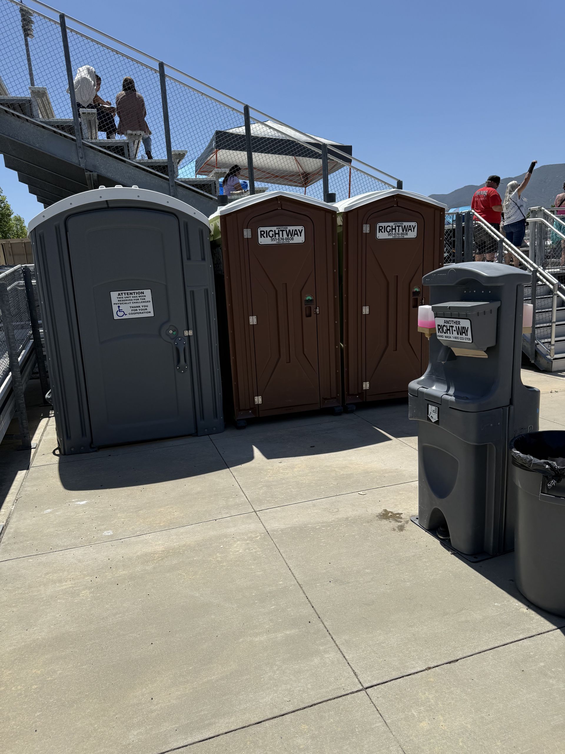 Three portable toilets and a hand sanitizer station on a concrete surface near bleachers.