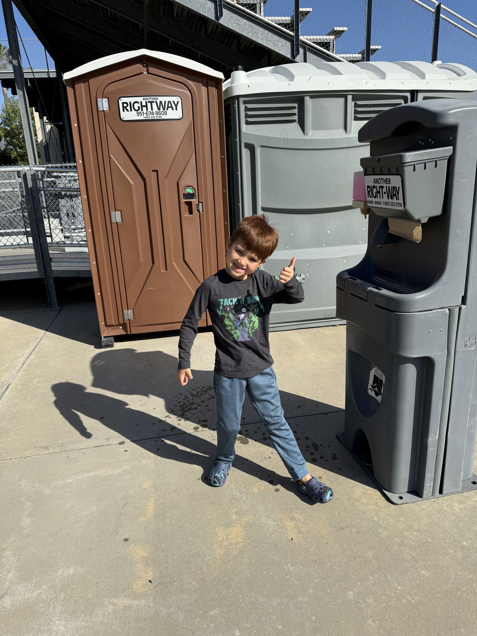 Young boy smiles and gives a thumbs-up gesture near portable restrooms outside on a sunny day.