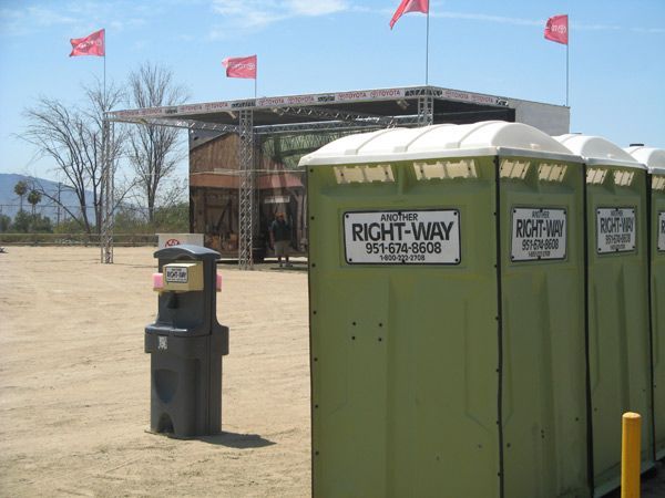 Row of green portable toilets in front of an outdoor stage with red flags in a dusty setting.