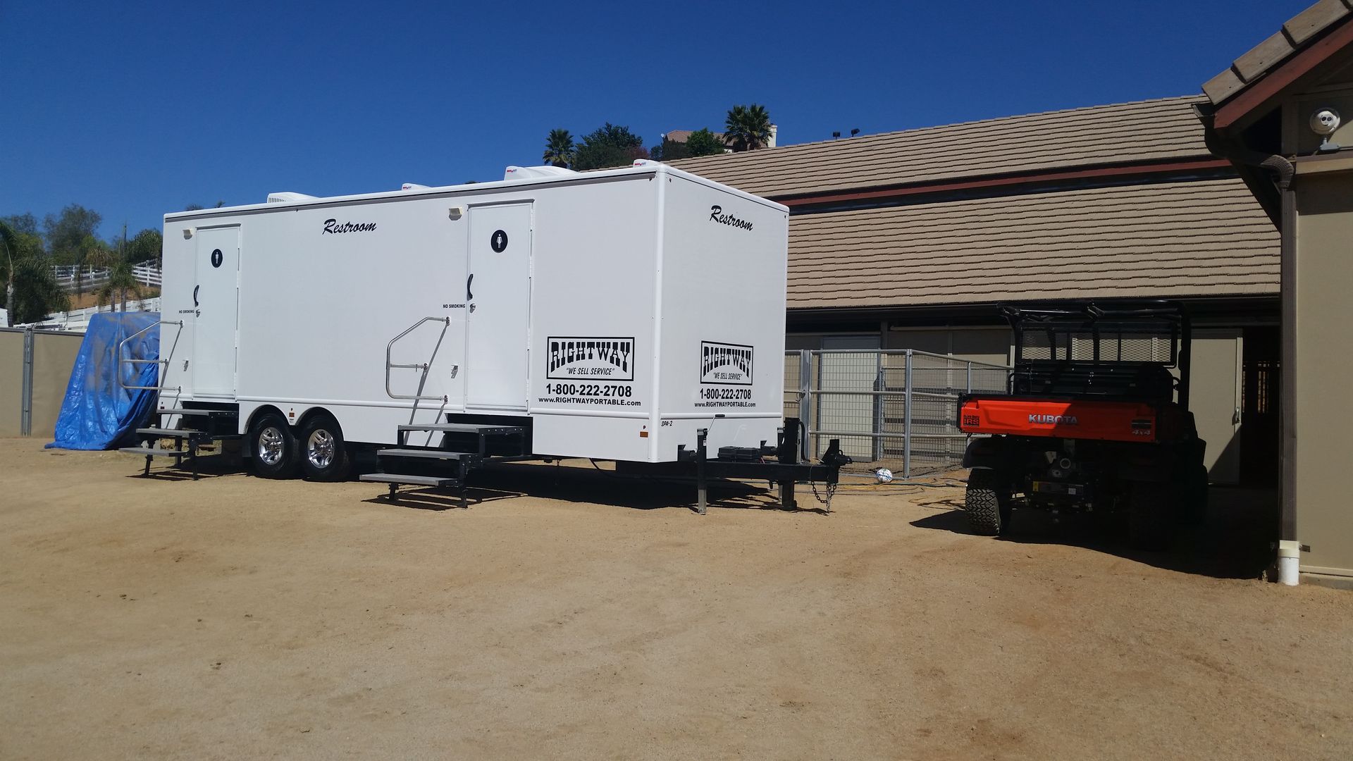 White portable restroom trailer next to a building. Orange utility vehicle parked nearby, blue tarp in background.