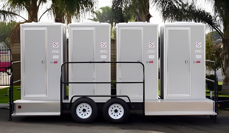 Four portable toilets on a trailer, gray, parked outdoors.