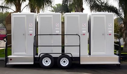 Four portable toilets on a trailer, gray, parked outdoors.