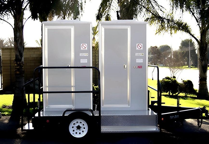 Two portable toilets on a trailer, gray in color, outdoors in a sunny environment.