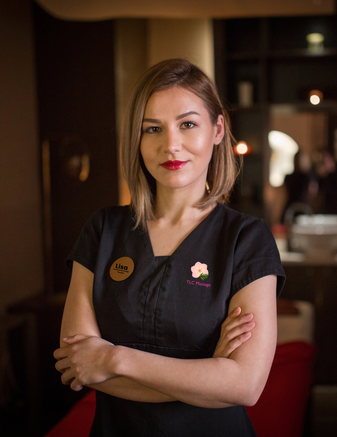 Apply Now! A female licensed massage therapist in a black shirt is standing with her arms crossed in a massage parlor