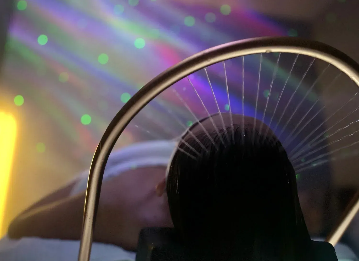 Person under a metal arch with strands, bathed in colorful lights, possibly receiving treatment.