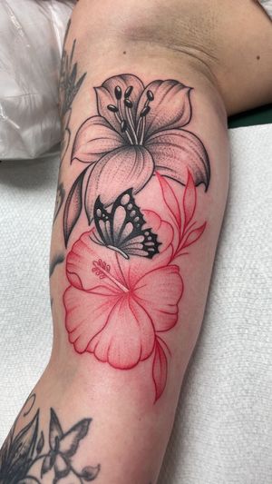 A black-ink lily sits above a red-ink hibiscus flower with a small butterfly resting on its petals, tattooed on an arm.