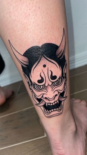 A black and grey tattoo of a Japanese Hannya mask on a person's lower leg.