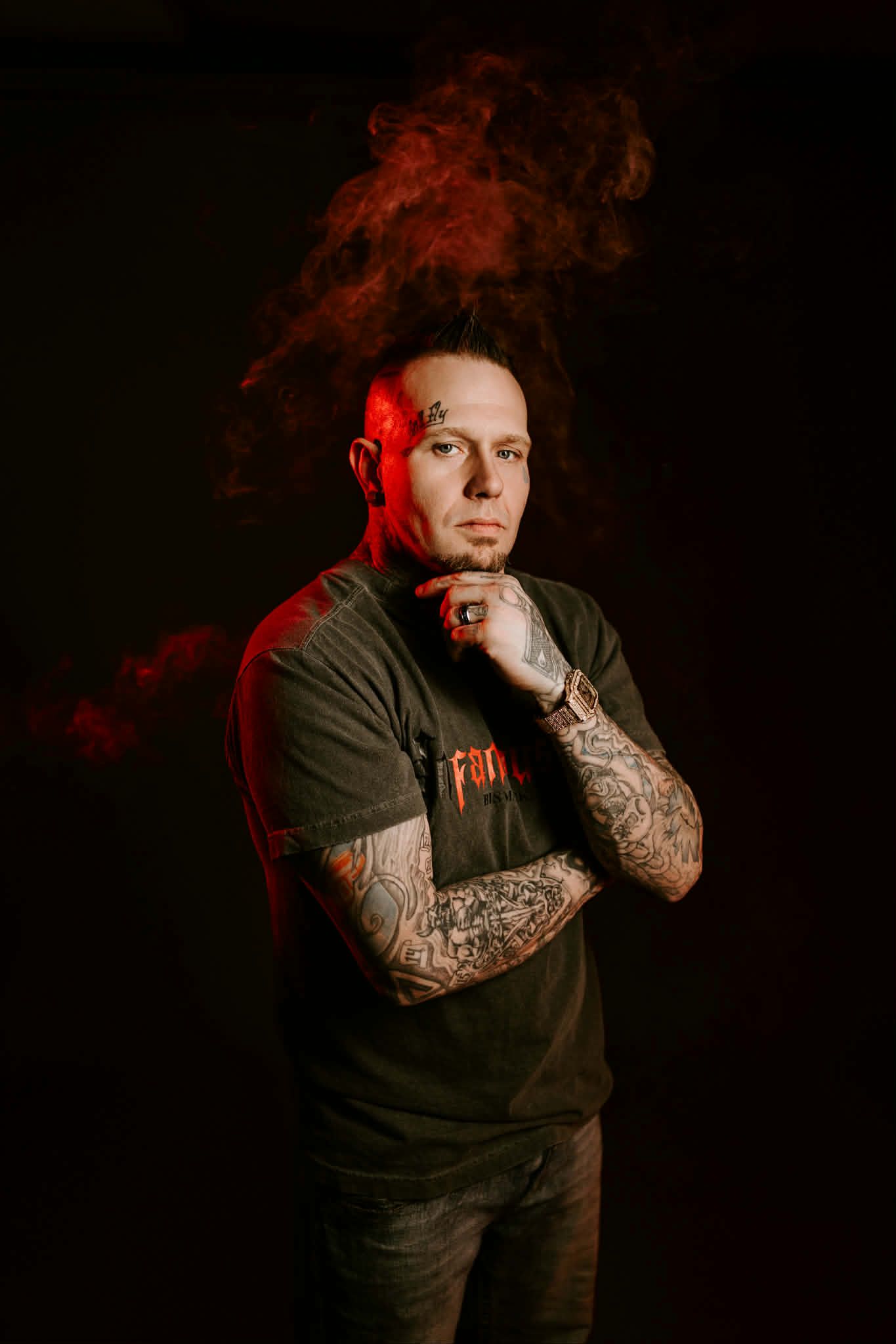 A person with arm tattoos and a face tattoo stands in dark red light with smoke rising around their head.