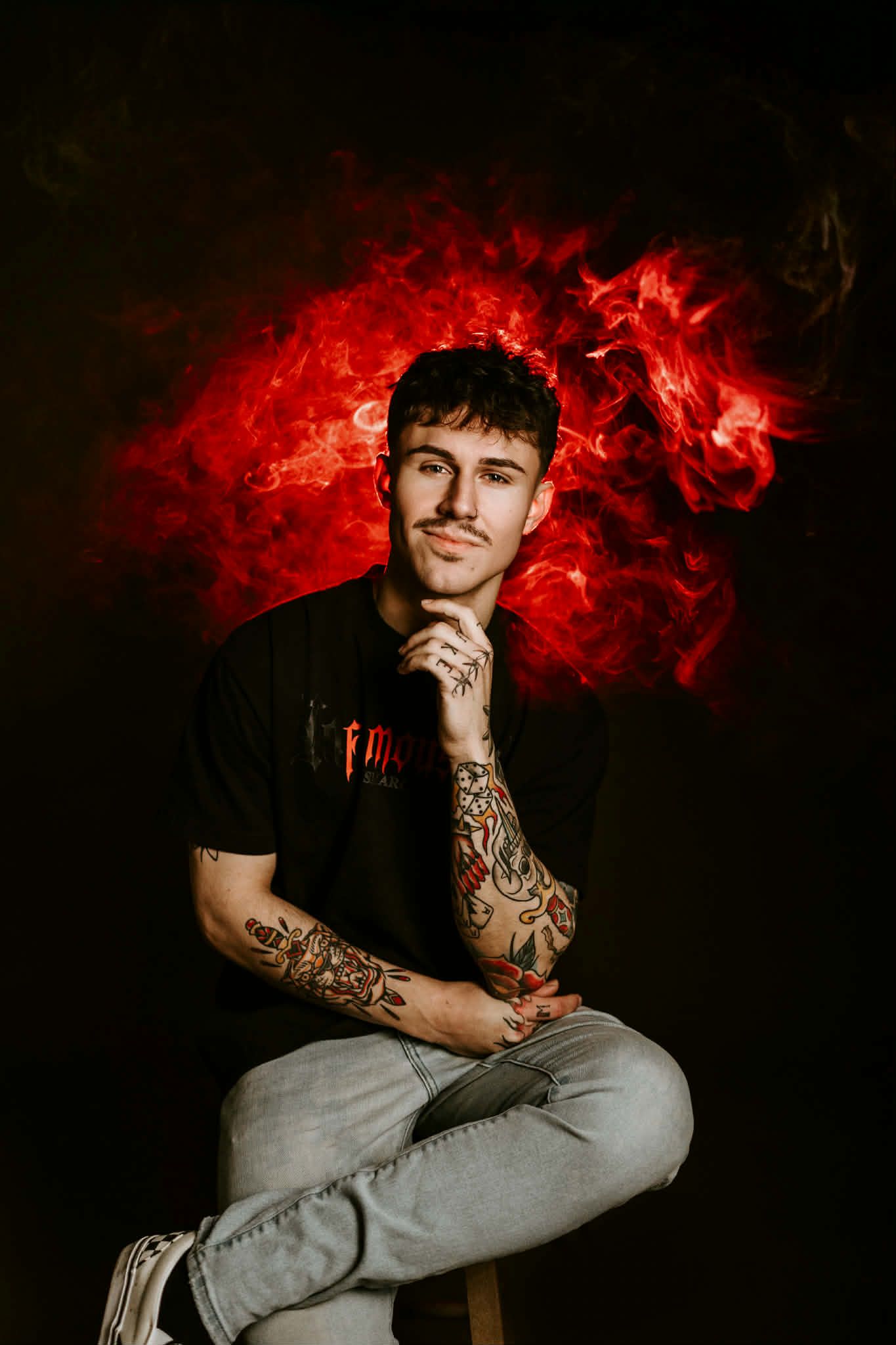A person with tattooed arms poses in a black T-shirt against a dark background with vibrant red smoke behind them.