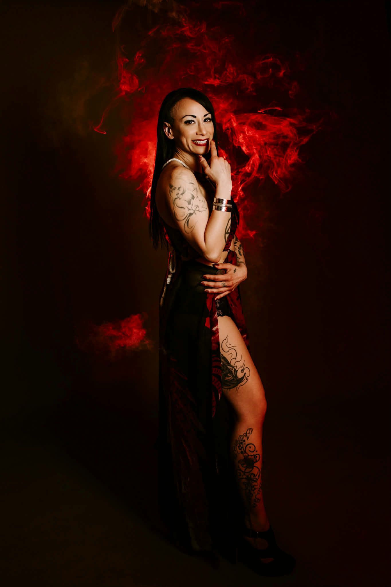 A person with visible tattoos stands against a dark, smoky red background, smiling and gesturing toward their face.