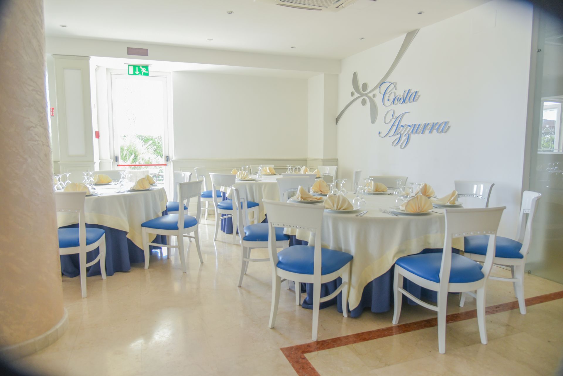 Luminosa sala da pranzo con tavoli rotondi apparecchiati per un evento formale, sedie blu e bianche e decorazioni alle pareti.
