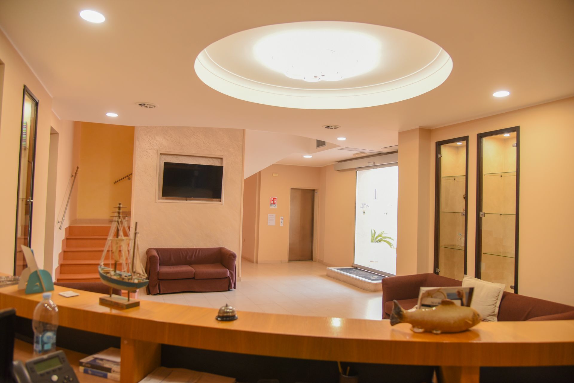 Hall d'albergo moderna con bancone della reception curvo, area lounge con divani, scalinata e lampadario circolare a soffitto.