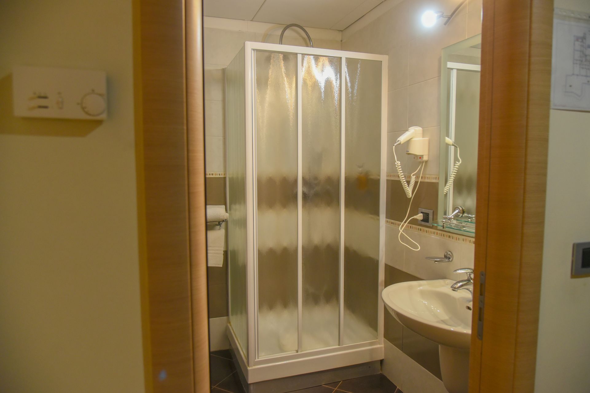 Piccolo bagno con box doccia bianco, lavandino, specchio e telaio della porta in legno.