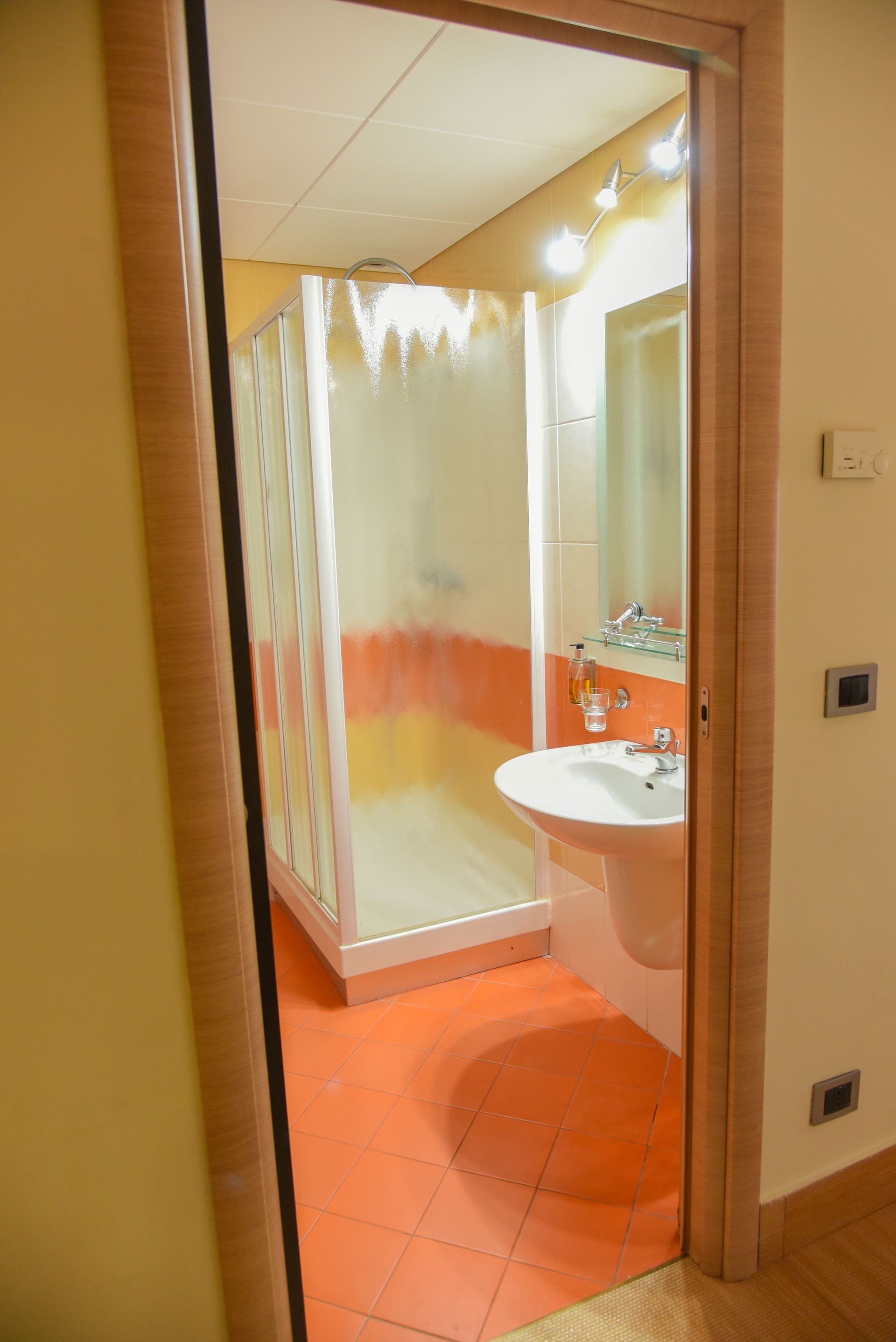 Ingresso del bagno con pavimento piastrellato arancione, lavabo bianco e box doccia satinato.