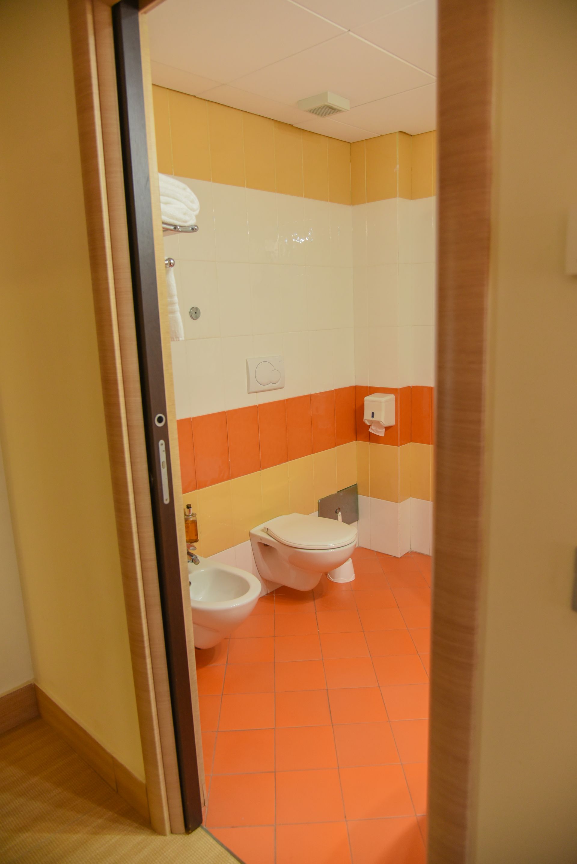 Piccolo bagno con pavimento e finiture in piastrelle arancioni, pareti bianche, lavandino, WC e porta aperta.