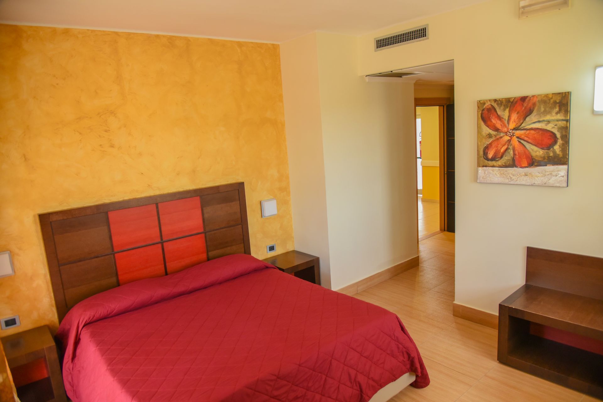 Camera da letto con copriletto rosso, mobili in legno, pareti gialle e una porta che conduce a un'altra stanza.