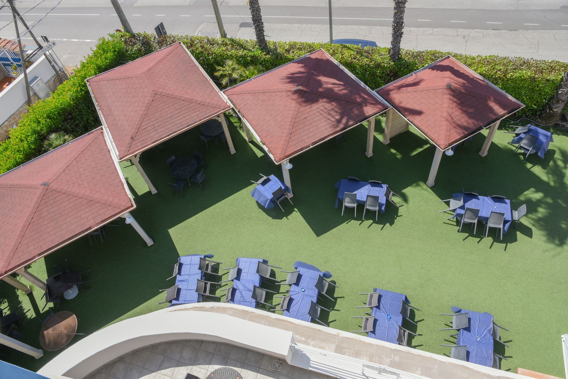 Patio esterno con gazebo dal tetto rosso, sedie blu e tavoli su un prato artificiale verde a bordo piscina.