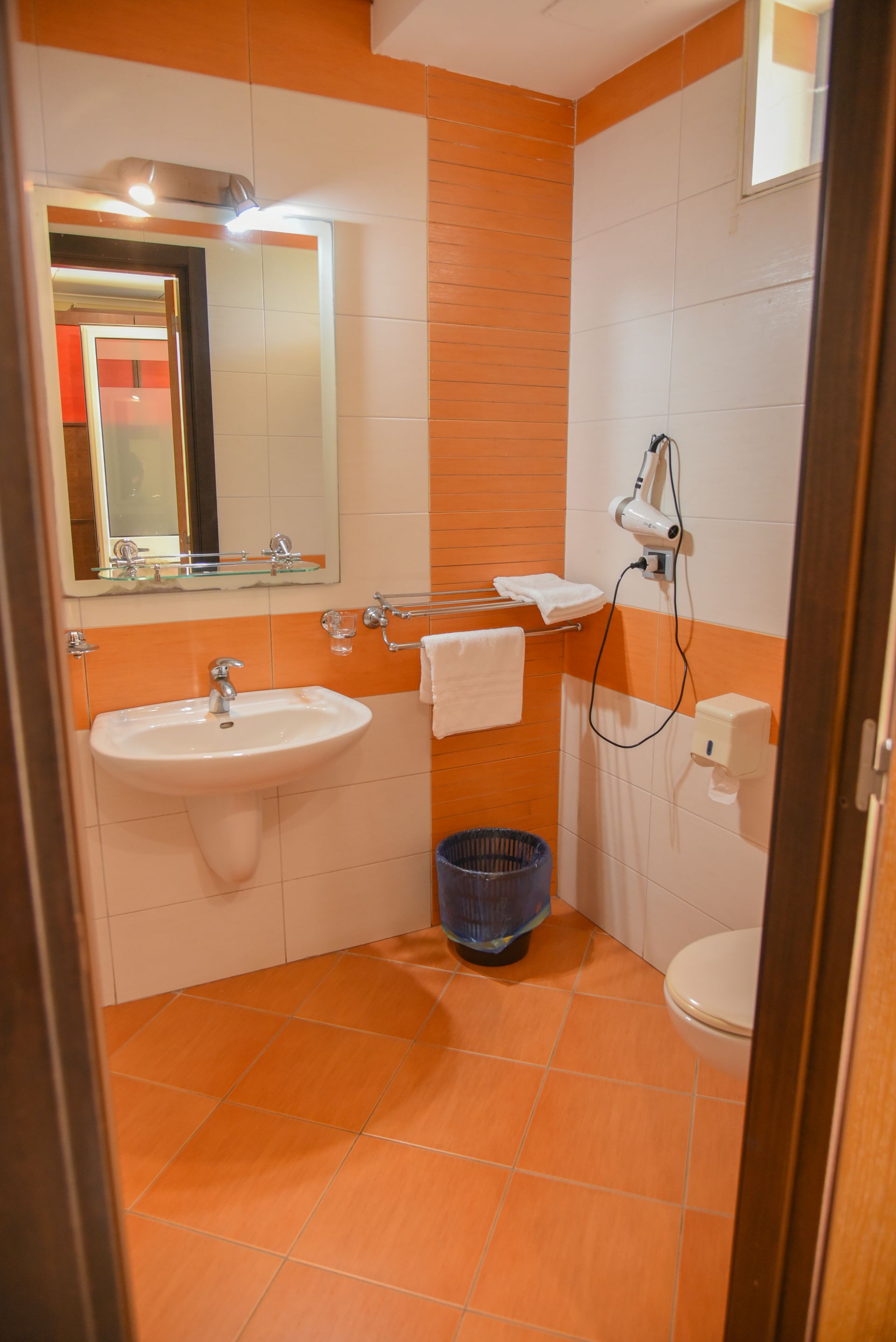 Piccolo bagno piastrellato di arancione con lavandino, specchio, WC, portasciugamani e doccetta.