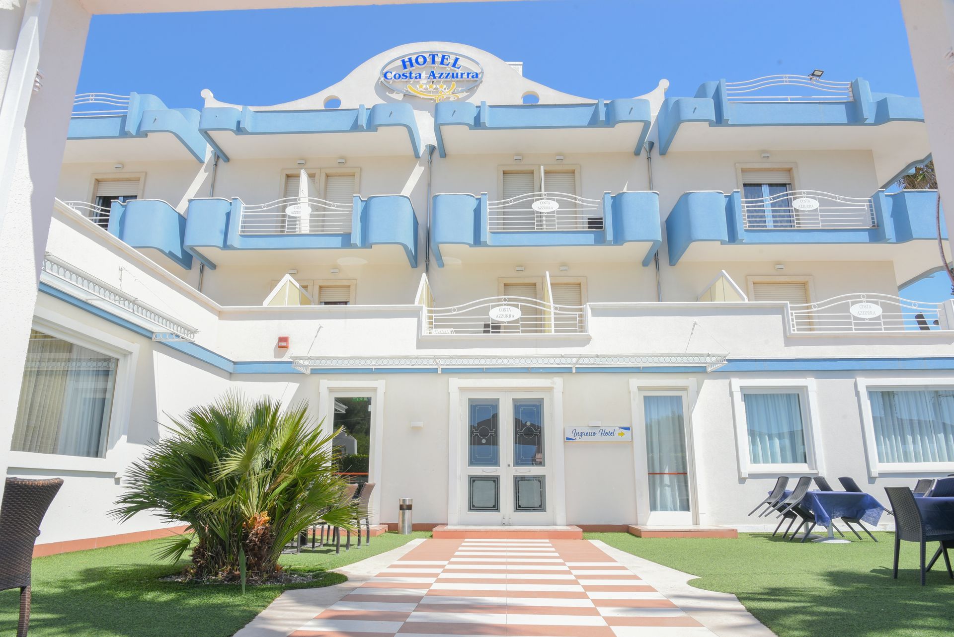 Hotel bianco sul mare con balconi, finiture blu e un vialetto a strisce che conduce all'ingresso.