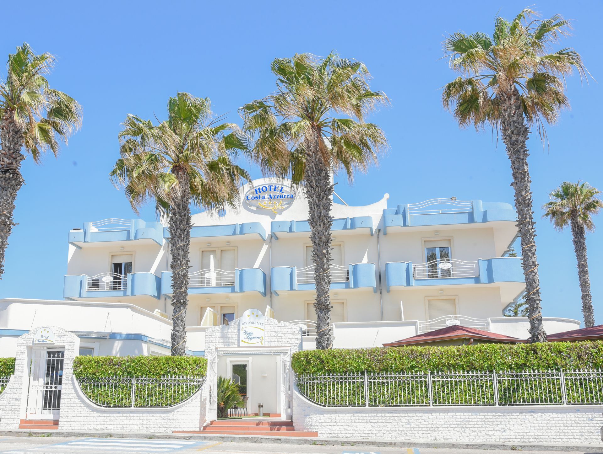 Hotel bianco sul mare con palme, balconi blu e un ingresso recintato sotto un cielo limpido