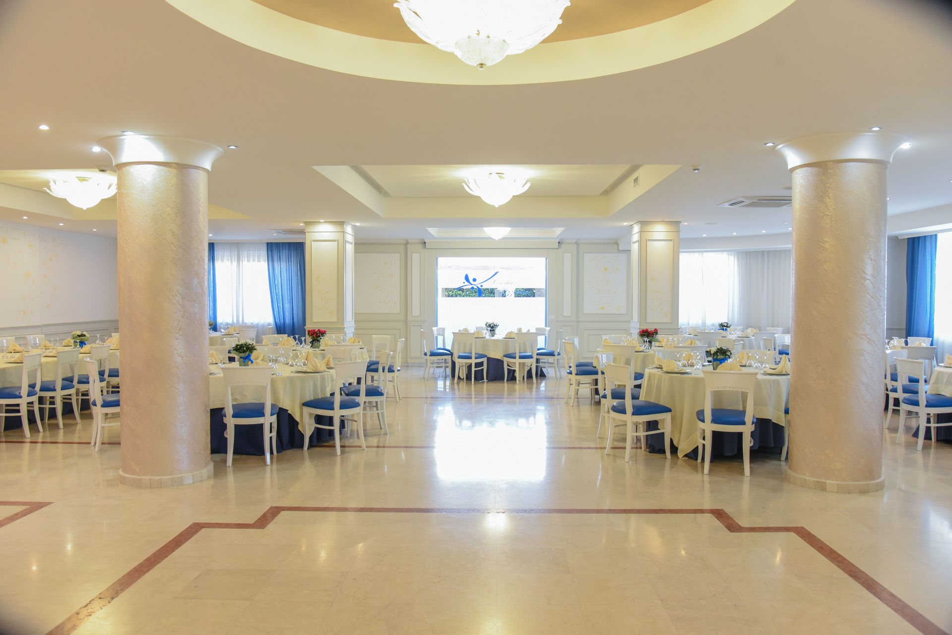 Luminosa sala per banchetti con tavoli rotondi, coprisedie bianchi e lampadari, vista dalla navata centrale.