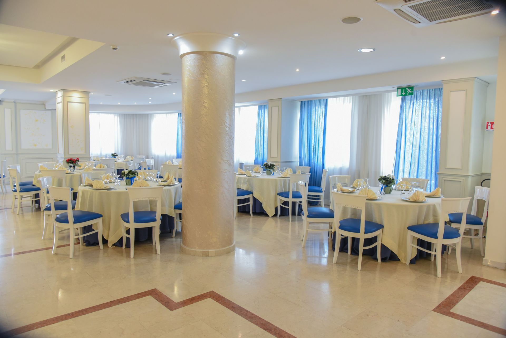 Luminosa sala da pranzo con tavoli rotondi, sedie blu e finestre a tutta altezza con tende blu.