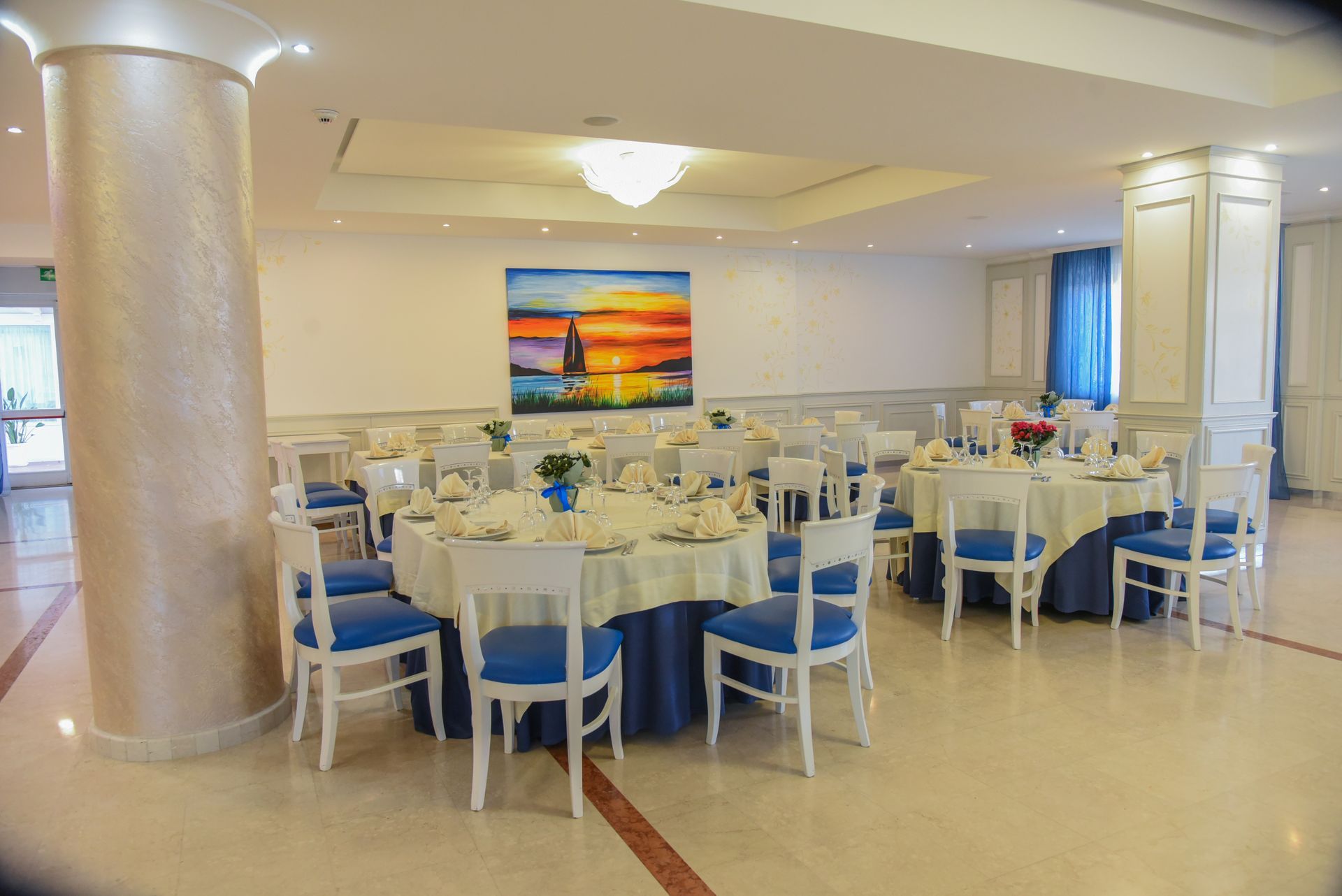 Elegante sala per banchetti con tavoli rotondi apparecchiati per un evento, sedie bianche e blu e un dipinto murale colorato.
