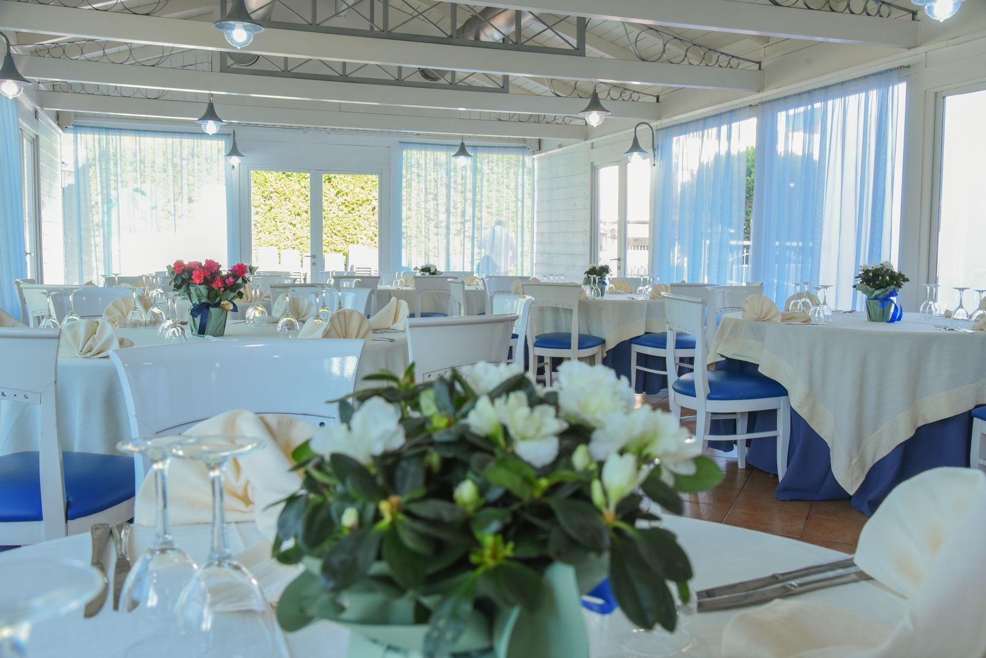 Luminosa sala per banchetti con tovaglie bianche, centrotavola floreali e grandi finestre affacciate sul mare.