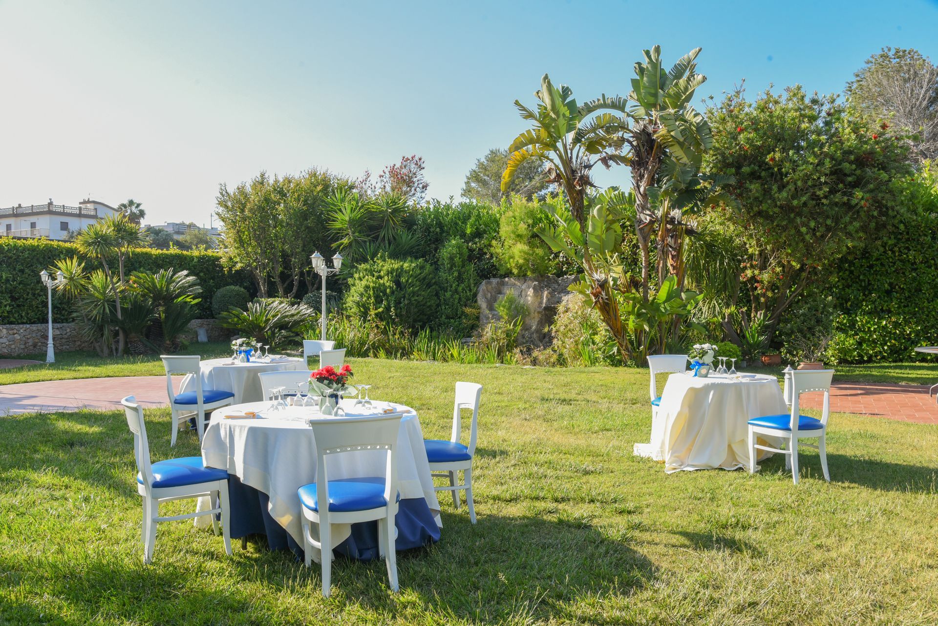 Giardino esterno con tavoli e sedie bianchi disposti su un prato erboso sotto alberi soleggiati