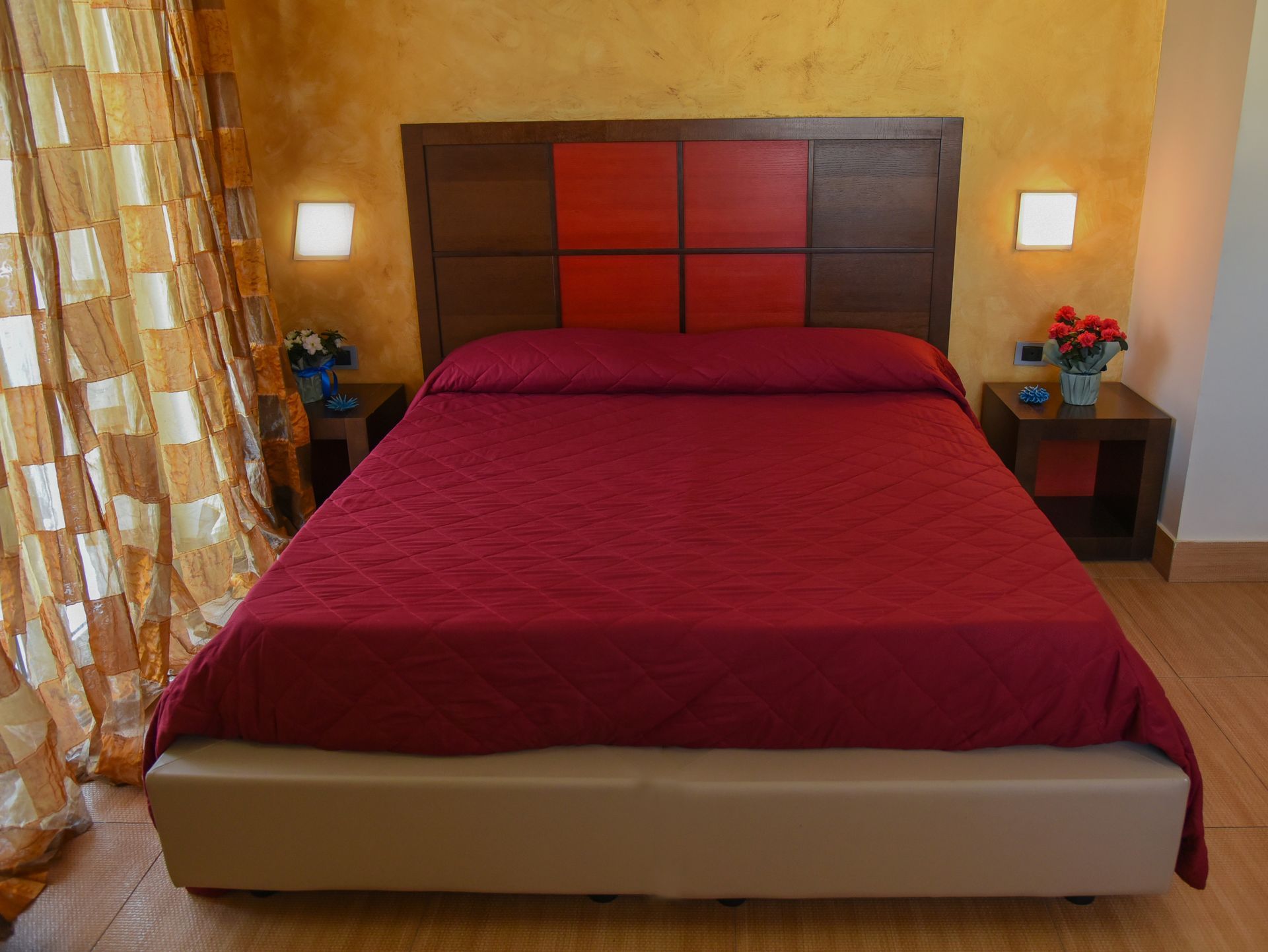 Camera da letto con copriletto rosso, testiera beige e pareti giallo caldo, fiancheggiata da piccoli comodini.