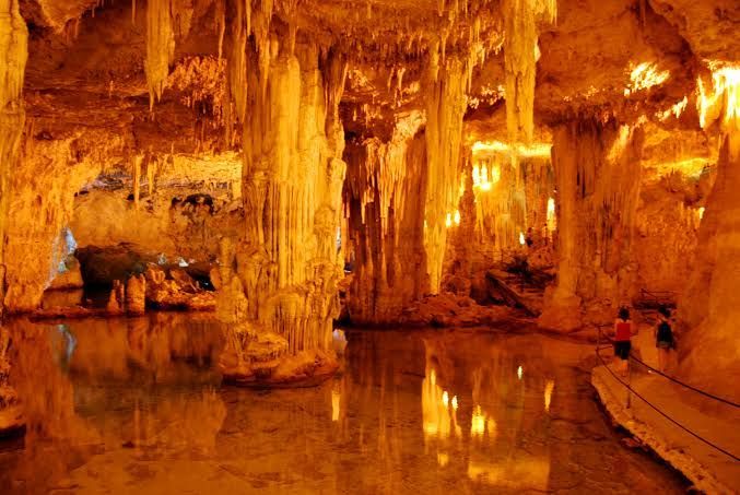 Interno della grotta illuminato da una luce arancione, con colonne di roccia, specchi d'acqua e una ringhiera che funge da passerella.