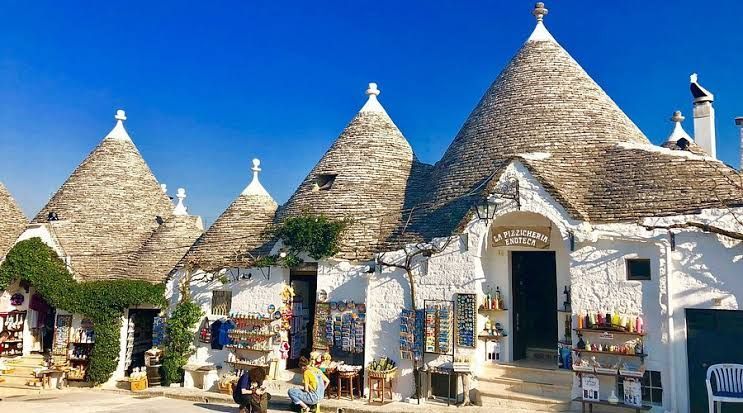 Edifici in pietra bianca in stile trullo con tetti conici sotto un cielo azzurro brillante, con persone fuori dai negozi.