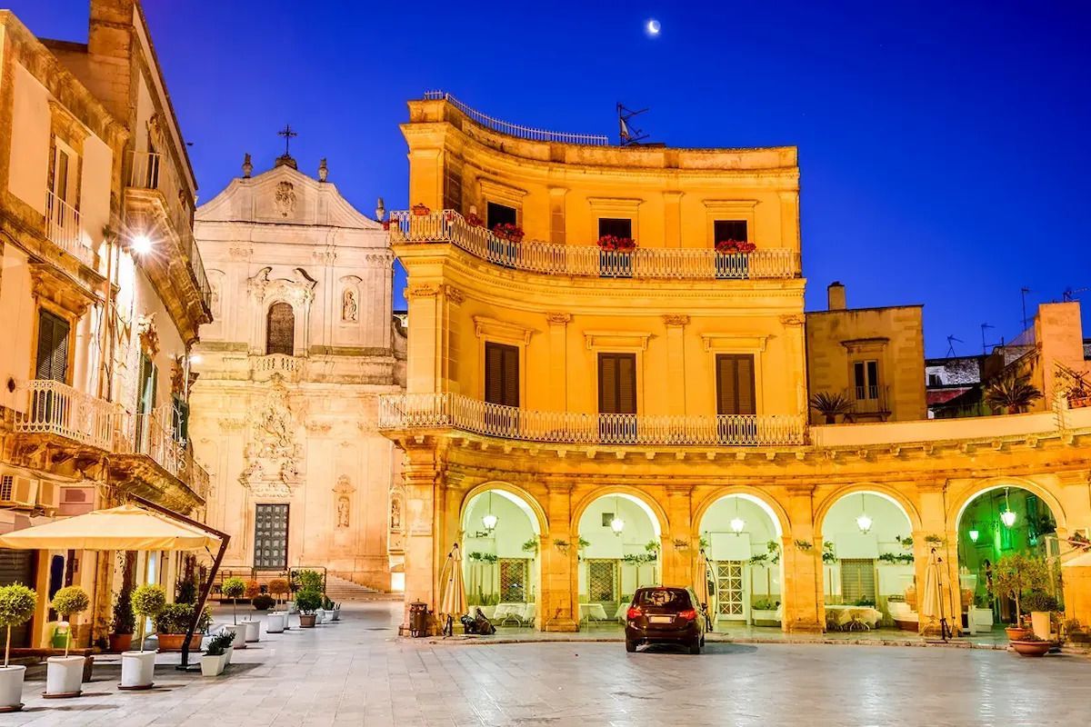 Piazza storica al crepuscolo con archi illuminati da una luce calda, edifici gialli e una piccola auto sotto un cielo blu intenso.