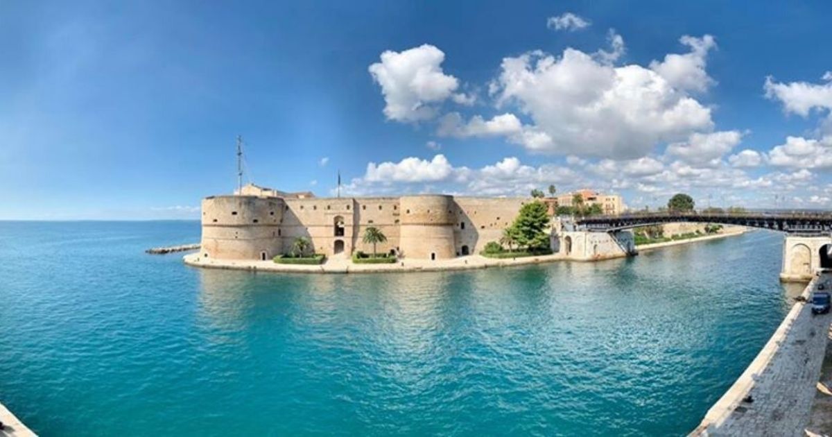 Fortezza in pietra sul mare, affacciata su una tranquilla baia turchese, sotto un cielo azzurro brillante con nuvole.