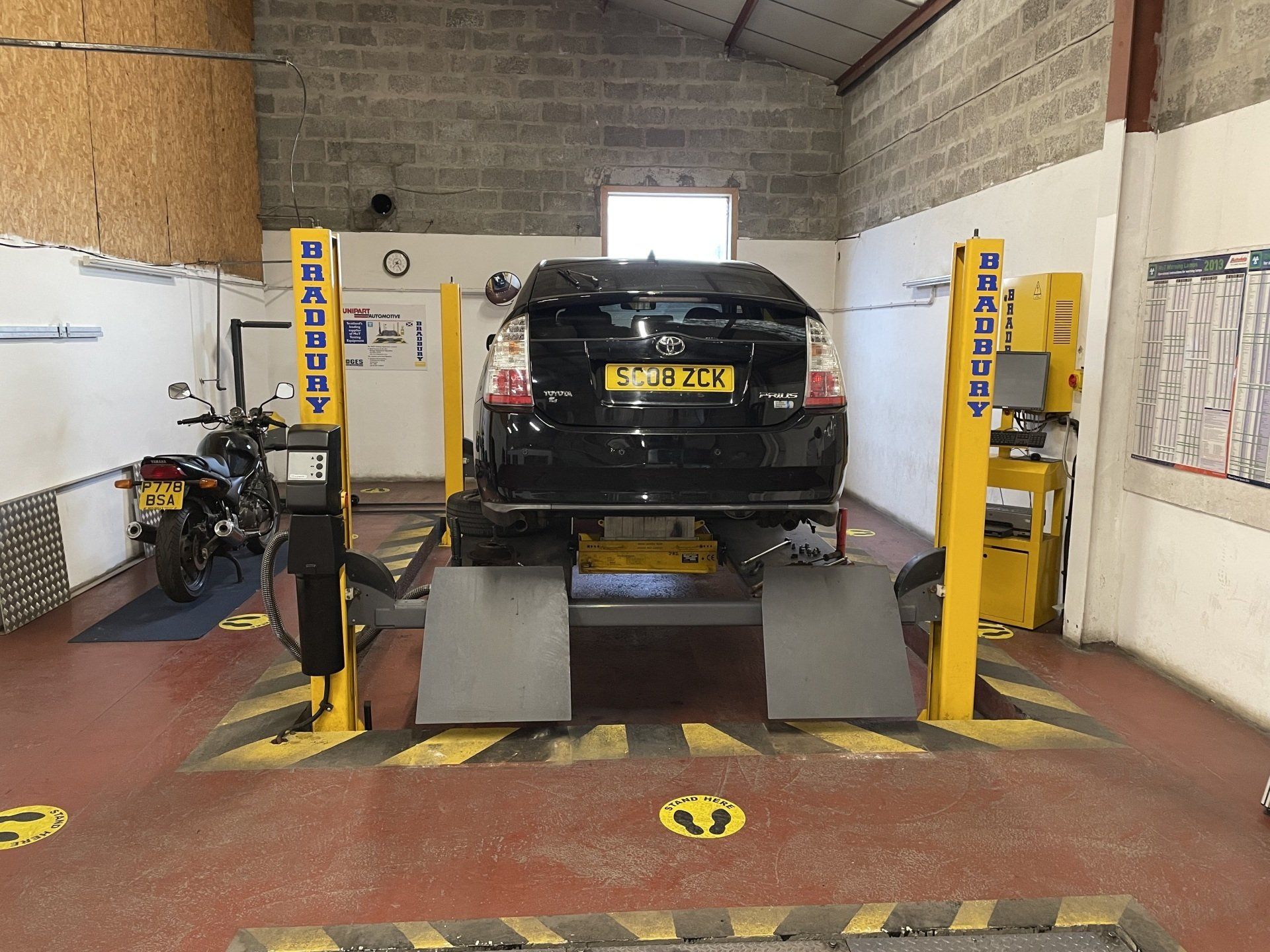 Black car on an alignment lift in an auto repair shop, with yellow equipment and gray ramps.