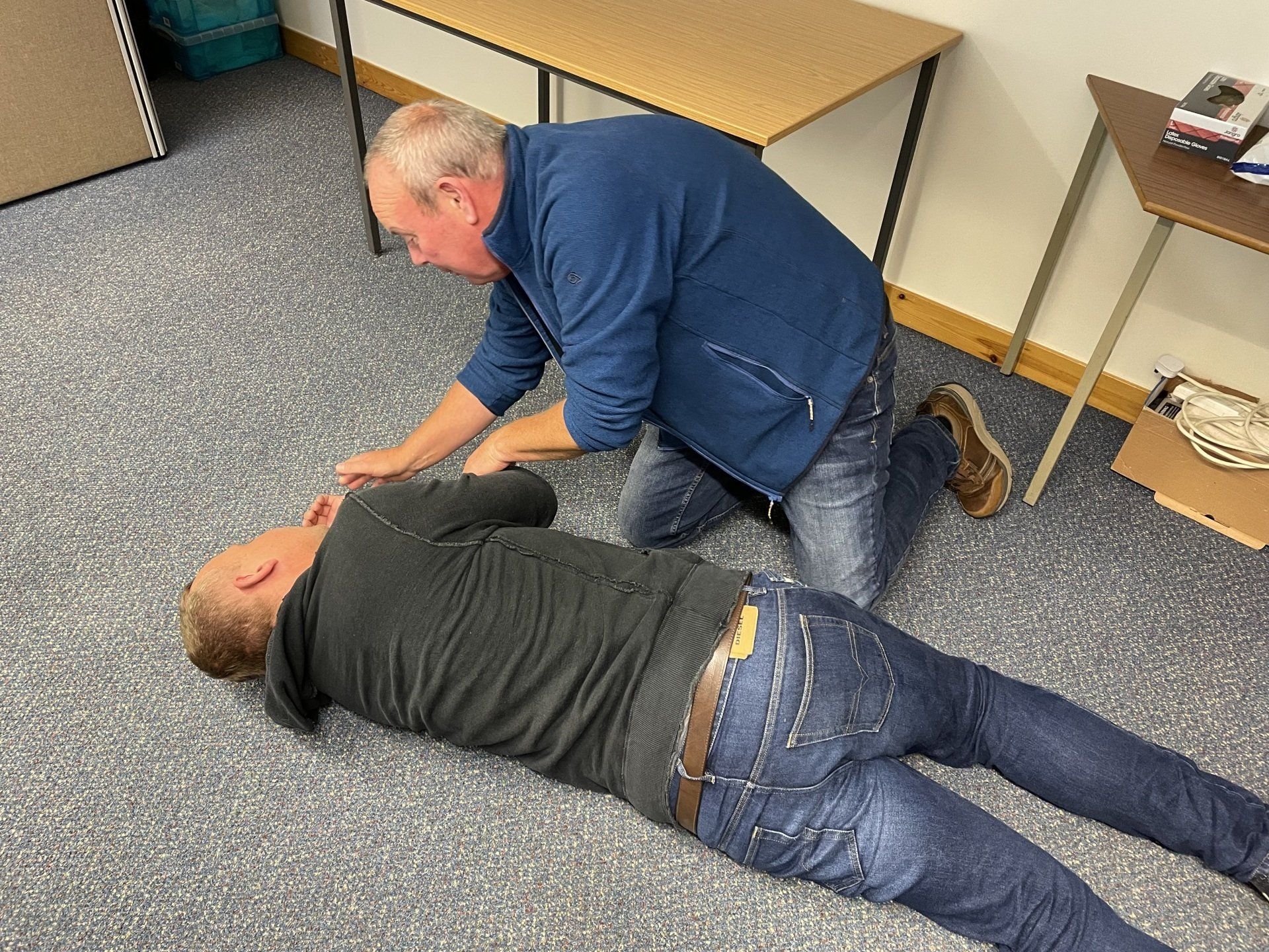 Two people on the floor, one kneeling and checking on the other in a room with tables.