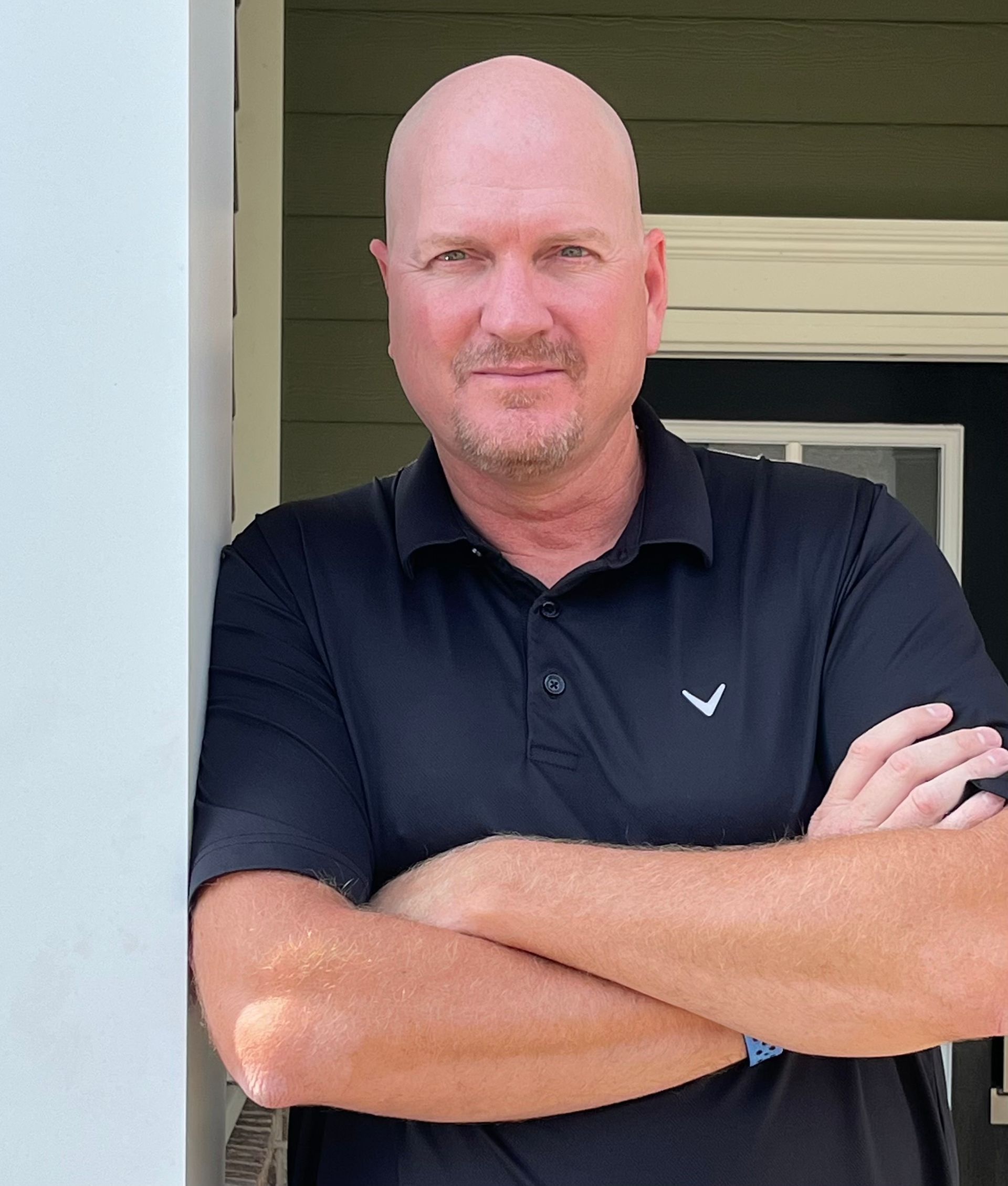 Bald man with arms crossed, wearing a black polo shirt, leaning against a white pillar, outside a green building.