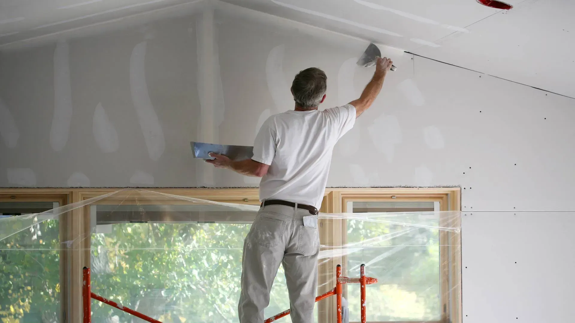 Worker drywalling a room, smoothing a white ceiling above windows and red scaffolding.