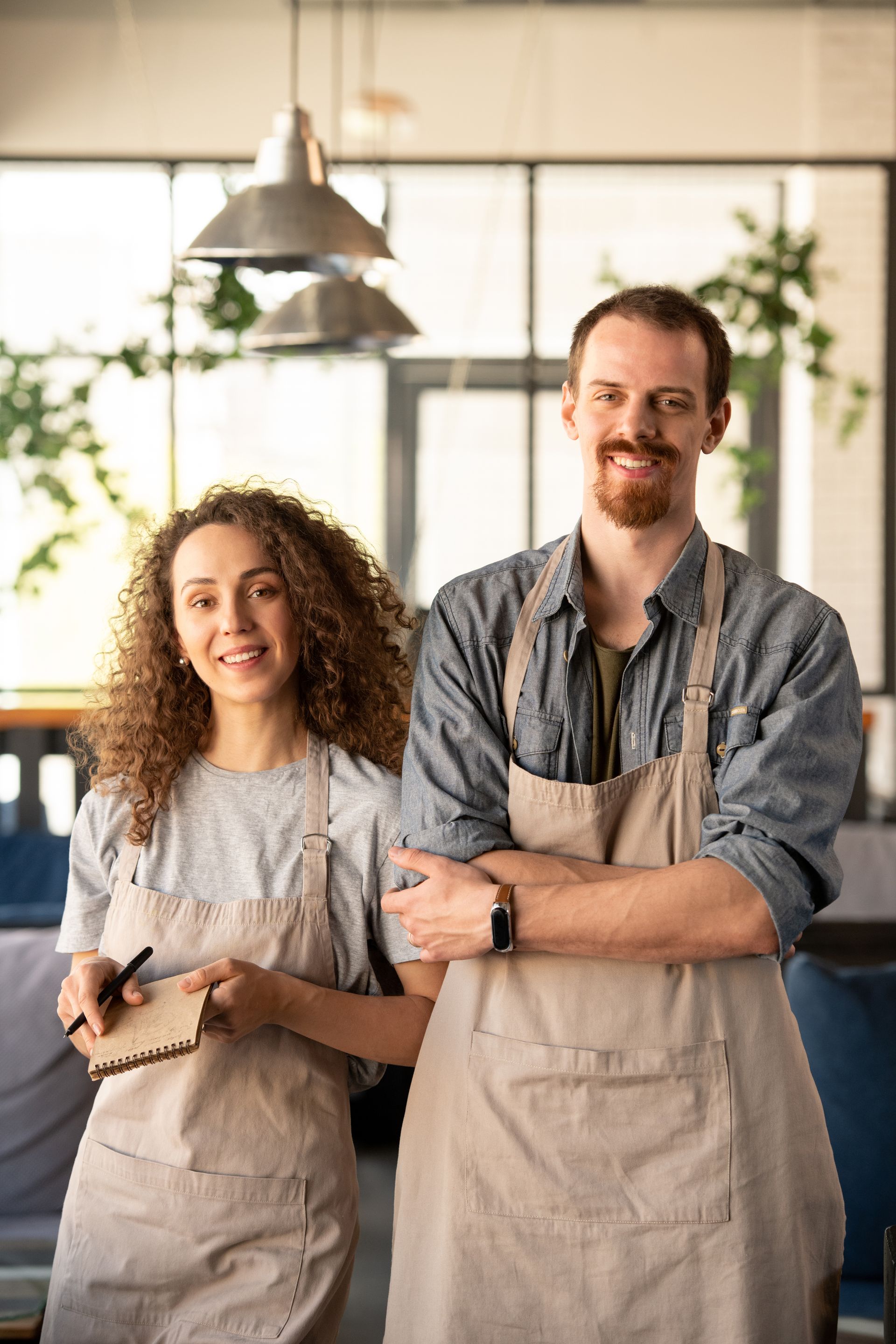 Two smiling café staff in beige aprons standing behind a counter in a bright kitchen