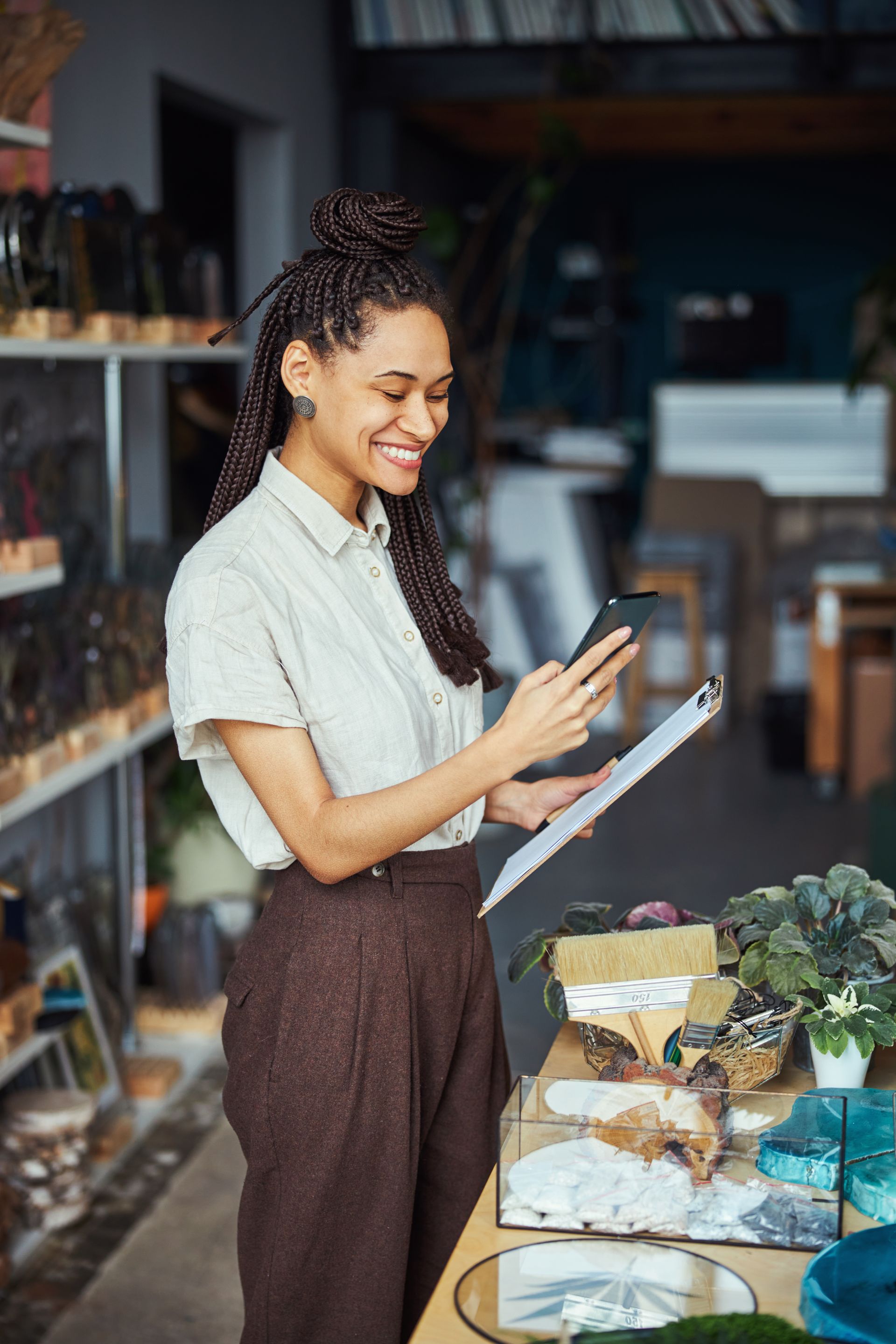 Person smiling and using a phone beside a display of flowers and baked goods in a shop