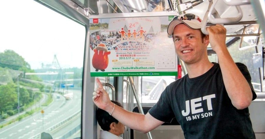 A smiling person wearing a JET t-shirt holds a handle and points to an advertisement displayed inside a train.