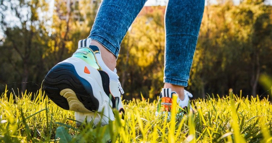 A person wearing blue jeans and white, chunky sneakers walks through a grassy field in bright, golden sunlight.