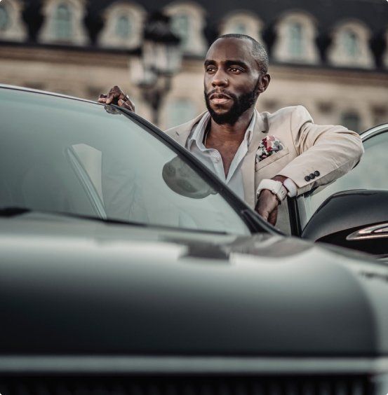 Man in beige suit leaning against a black car, looking off to the side. Exterior setting with buildings.