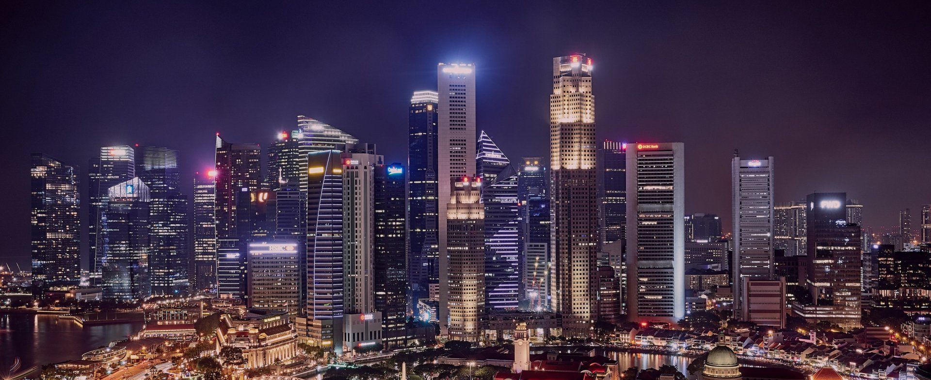 Nighttime cityscape of lit skyscrapers against a dark purple sky. City lights twinkle below.