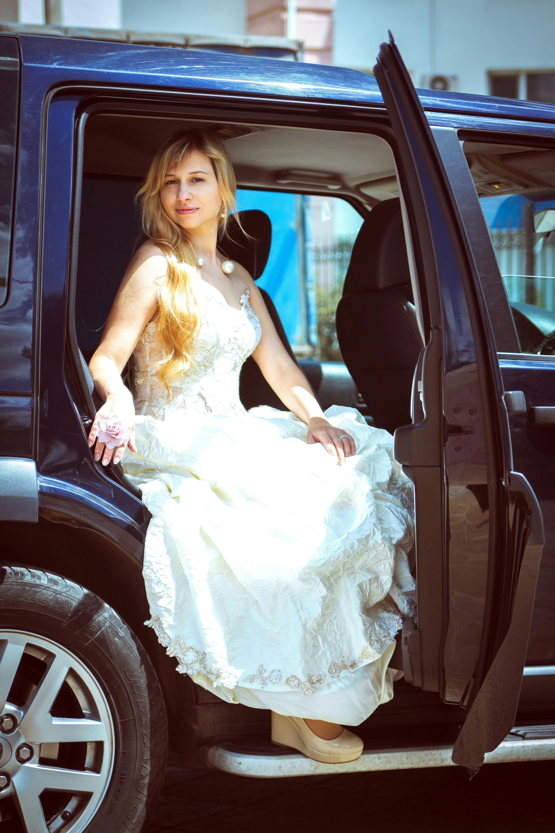 Woman in a white gown exiting a dark blue SUV. She is smiling.