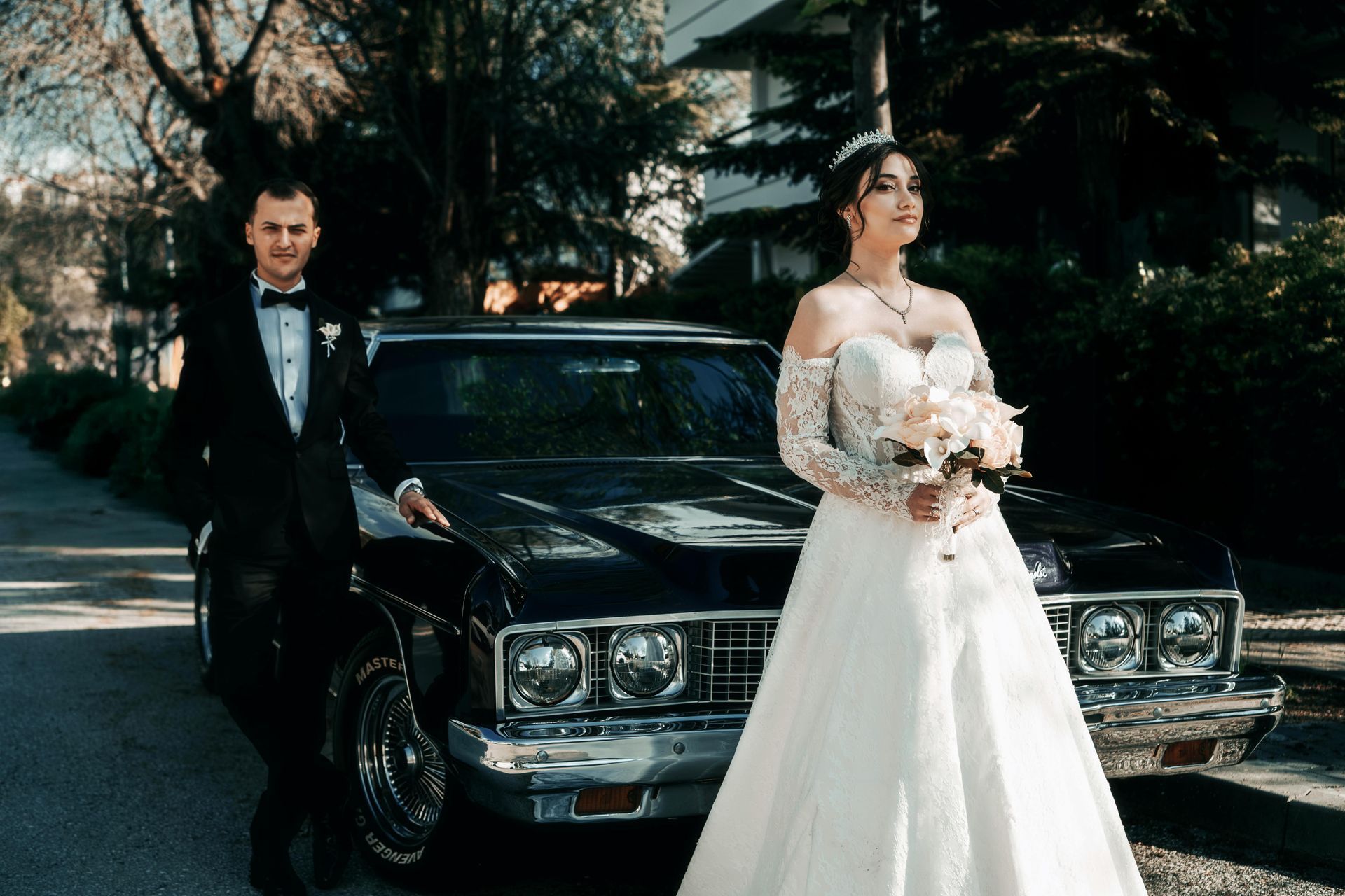 Bride and groom pose by a black classic car on a tree-lined street. The bride holds flowers.