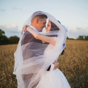 Bride and groom embrace, veiled by a sheer white veil, in a wheat field. They are looking at each other romantically.