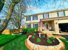 Two-story beige house with green shutters and landscaping, American flag.