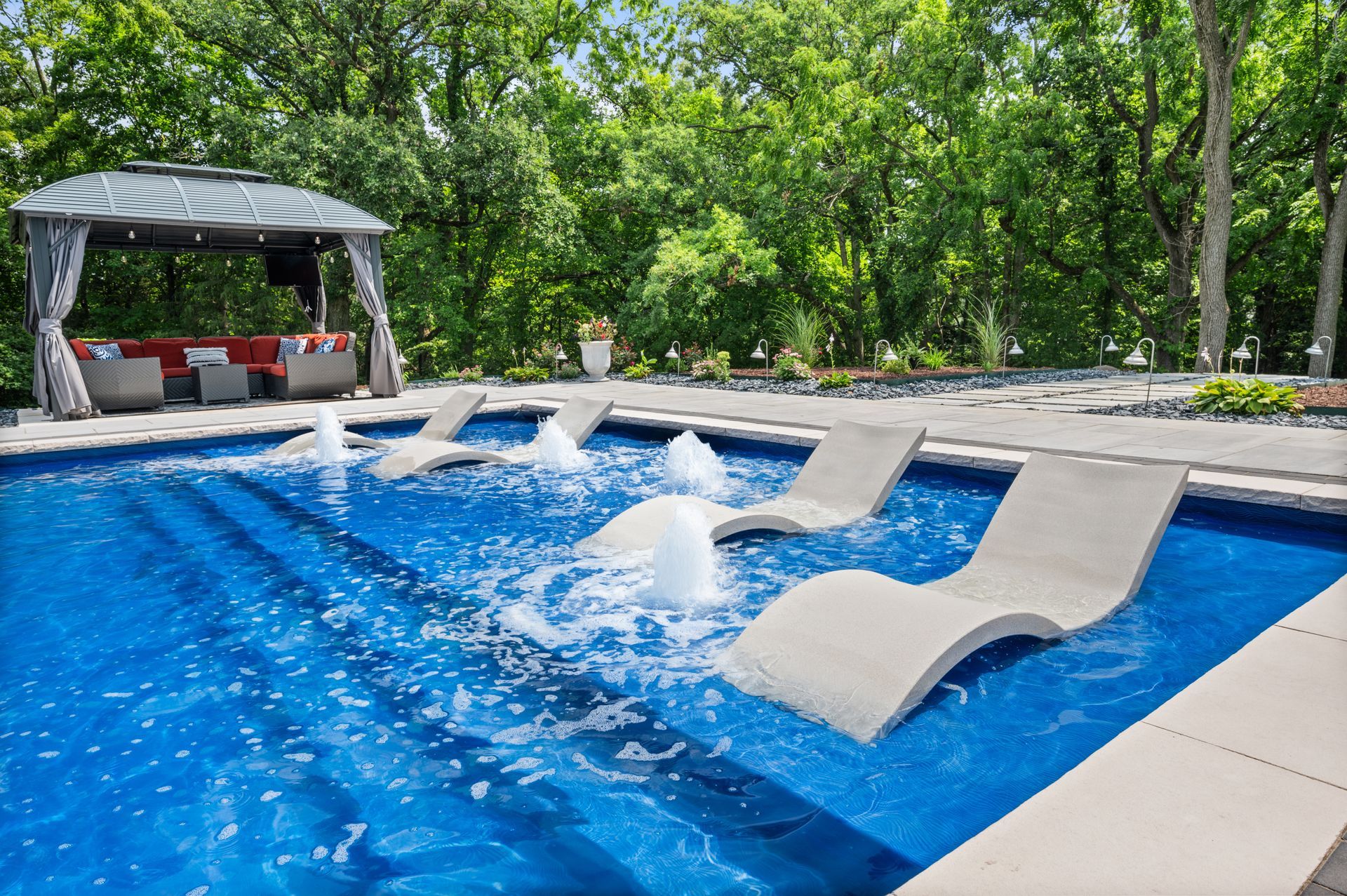Backyard pool with a splash near lounge chairs and a shaded gazebo amid trees